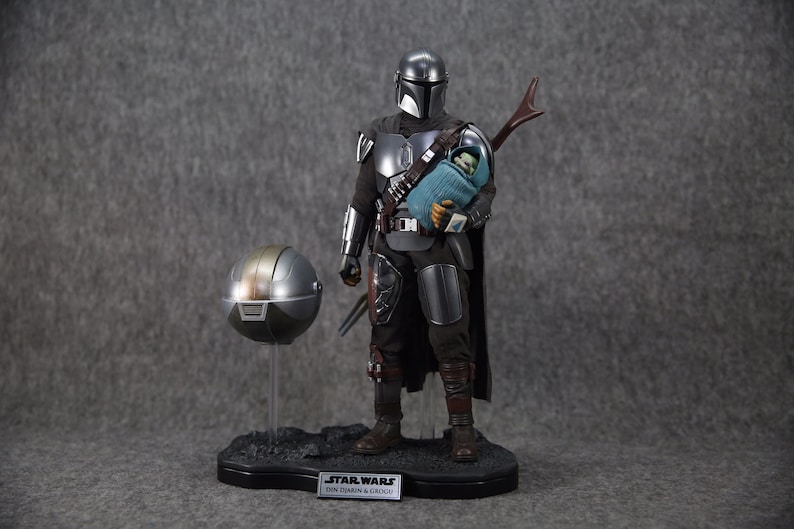 Puede incluir: Figura detallada de The Mandalorian sosteniendo a Grogu, con un casco separado en un soporte. El Mandaloriano viste una armadura plateada y negra, sosteniendo a Grogu envuelto en una manta azul. La base tiene el logo de Star Wars y texto.