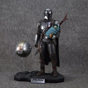 Puede incluir: Figura detallada de The Mandalorian sosteniendo a Grogu, con un casco separado en un soporte. El Mandaloriano viste una armadura plateada y negra, sosteniendo a Grogu envuelto en una manta azul. La base tiene el logo de Star Wars y texto.