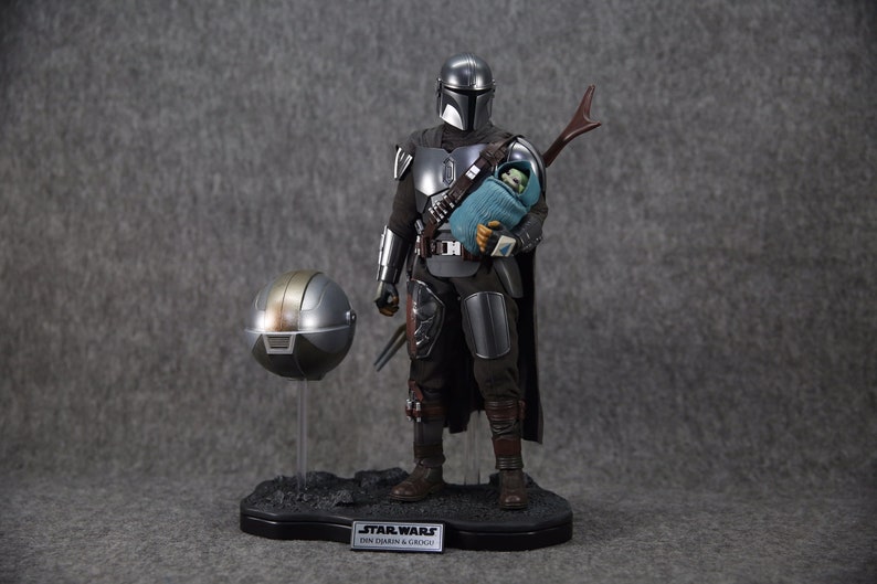Puede incluir: Figura detallada de The Mandalorian sosteniendo a Grogu, con un casco separado en un soporte. El Mandaloriano viste una armadura plateada y negra, sosteniendo a Grogu envuelto en una manta azul. La base tiene el logo de Star Wars y texto.