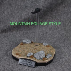 May include: A clear plastic display stand with a black shoe holder. The base is a brown and gray textured surface with rocks and green foliage. The base has a silver nameplate that says "Movie Title Character Name".