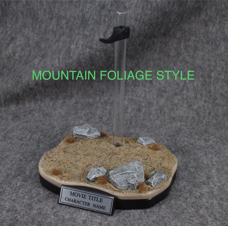 May include: A clear plastic display stand with a black shoe holder. The base is a brown and gray textured surface with rocks and green foliage. The base has a silver nameplate that says "Movie Title Character Name".