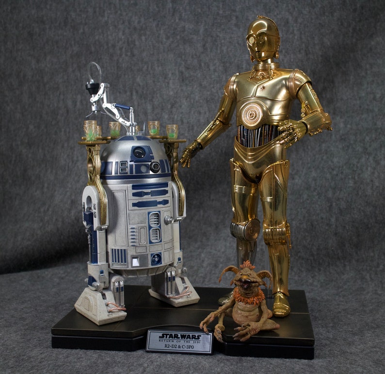 May include: A Star Wars collectible set featuring R2-D2 and C-3PO. R2-D2 is a blue and white droid with a gold serving tray. C-3PO is a gold droid with a silver head. The set includes a small brown creature with a red scarf.
