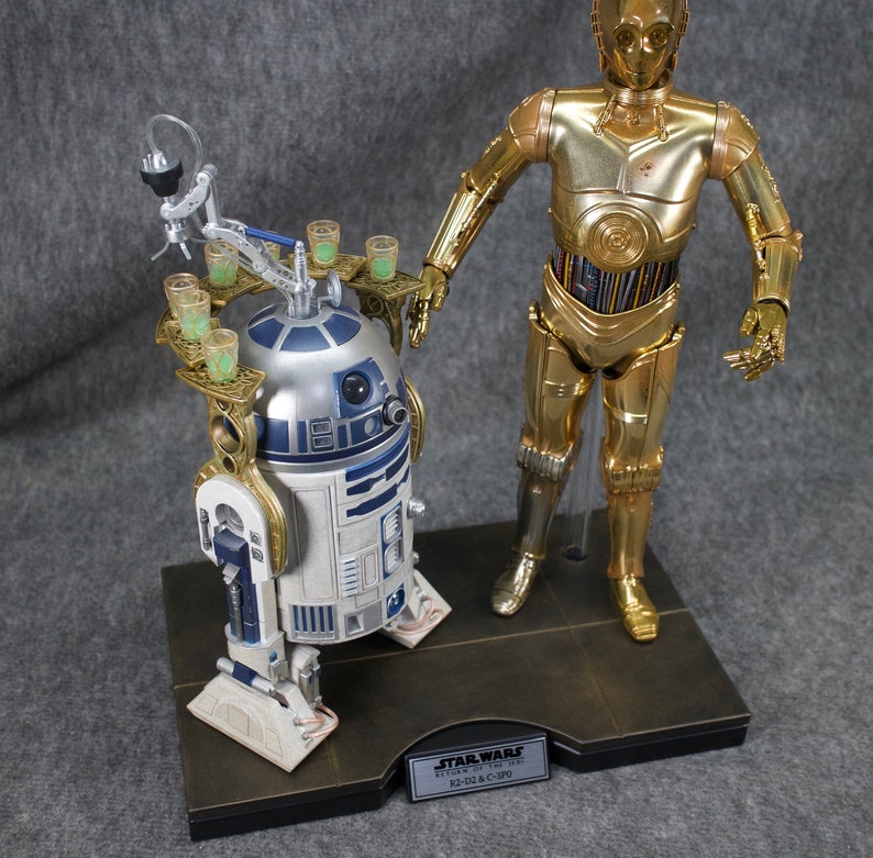 May include: A gold-colored C-3PO Star Wars action figure stands next to a blue and white R2-D2 action figure. Both figures are on a black base with a gold trim. The base has a plaque that reads "STAR WARS Episode IV: A New Hope R2-D2 & C-3PO".