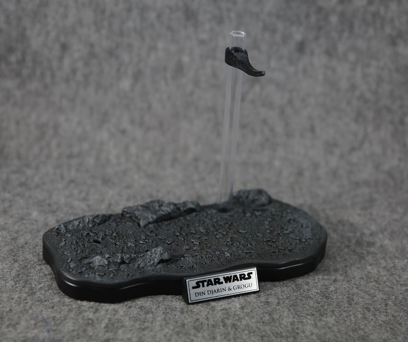 Puede incluir: Un soporte de exhibici&oacute;n negro para figuras de acci&oacute;n con una base texturizada y una varilla de soporte transparente. El soporte presenta un peque&ntilde;o reposapi&eacute;s negro y una placa rectangular con el texto "STAR WARS DIN DJARIN & GROGU".