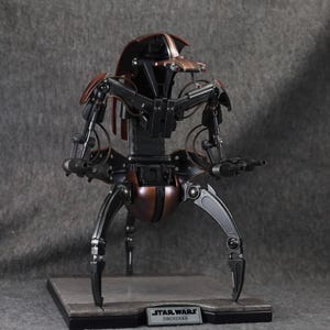 Puede incluir: Modelo detallado de un Droideka de Star Wars, un droide destructor rodante. La figura es principalmente negra y bronce, con extremidades y armas articuladas. Se encuentra sobre una base rectangular con la etiqueta "Star Wars Droideka".