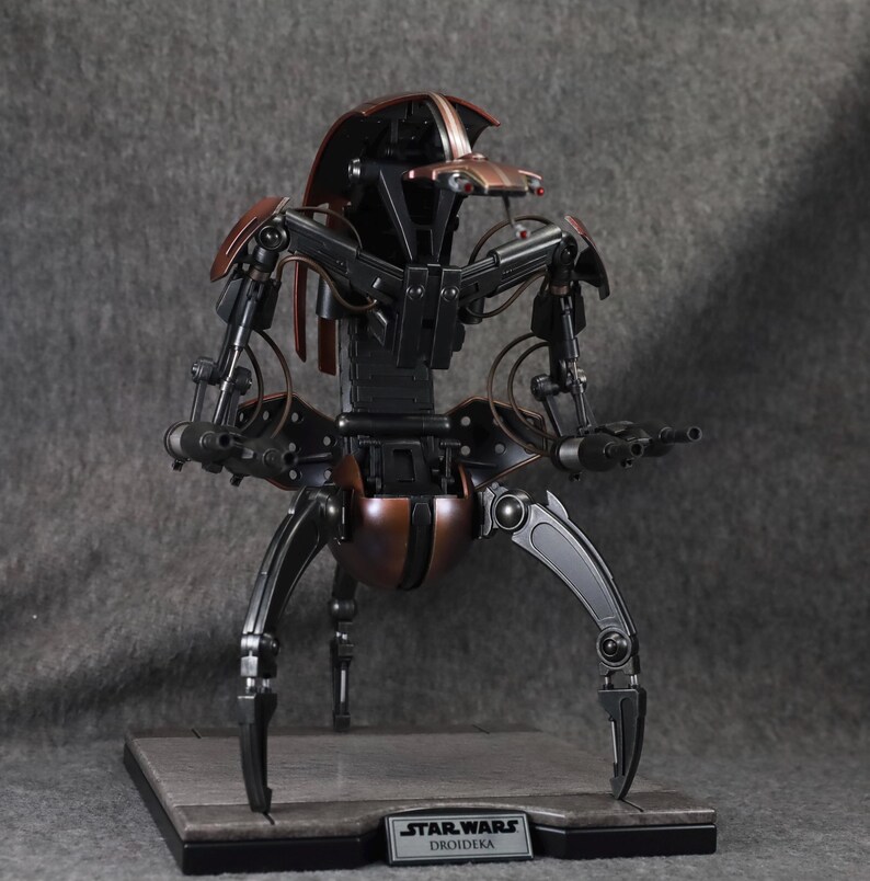 Puede incluir: Modelo detallado de un Droideka de Star Wars, un droide destructor rodante. La figura es principalmente negra y bronce, con extremidades y armas articuladas. Se encuentra sobre una base rectangular con la etiqueta "Star Wars Droideka".
