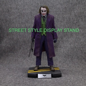 K&ouml;nnte beinhalten: Eine lila und gr&uuml;ne Joker-Actionfigur mit einer Pistole und einem Ausstellungsst&auml;nder. Der St&auml;nder hat eine schwarze Basis mit gelben Streifen und eine schwarze Plakette mit der Aufschrift "The Dark Knight Joker".
