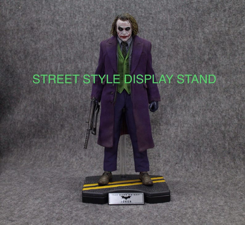 K&ouml;nnte beinhalten: Eine lila und gr&uuml;ne Joker-Actionfigur mit einer Pistole und einem Ausstellungsst&auml;nder. Der St&auml;nder hat eine schwarze Basis mit gelben Streifen und eine schwarze Plakette mit der Aufschrift "The Dark Knight Joker".