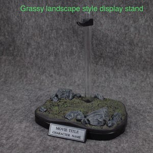 May include: A black display stand with a grassy landscape base. The stand has a clear acrylic rod and a black base with a label that says "Movie Title" and "Character Name".