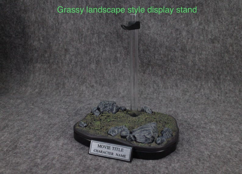 May include: A black display stand with a grassy landscape base. The stand has a clear acrylic rod and a black base with a label that says "Movie Title" and "Character Name".