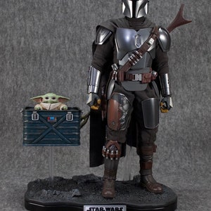 Puede incluir: Figura de acci&oacute;n detallada de The Mandalorian, Din Djarin, con armadura plateada y negra, con una figura de Grogu en una caja azul. Las figuras est&aacute;n sobre una base negra con el logo de Star Wars y los nombres de los personajes.
