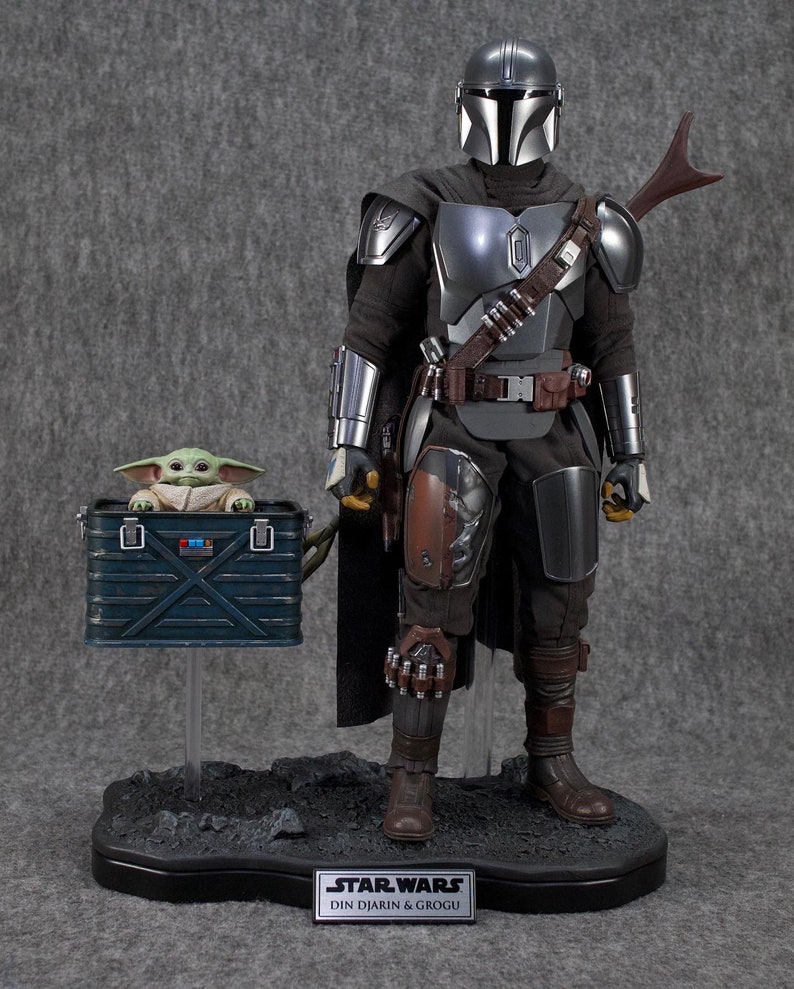 Puede incluir: Figura de acci&oacute;n detallada de The Mandalorian, Din Djarin, con armadura plateada y negra, con una figura de Grogu en una caja azul. Las figuras est&aacute;n sobre una base negra con el logo de Star Wars y los nombres de los personajes.