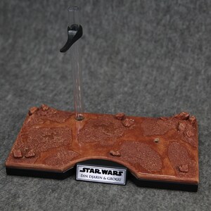 May include: A brown and black display stand with a textured surface that resembles a desert landscape. The stand has a clear plastic rod with a black handle and a silver plaque that reads "STAR WARS DIN DJARIN & GROGU".