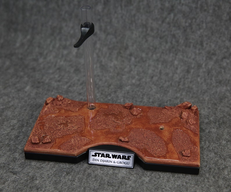May include: A brown and black display stand with a textured surface that resembles a desert landscape. The stand has a clear plastic rod with a black handle and a silver plaque that reads "STAR WARS DIN DJARIN & GROGU".