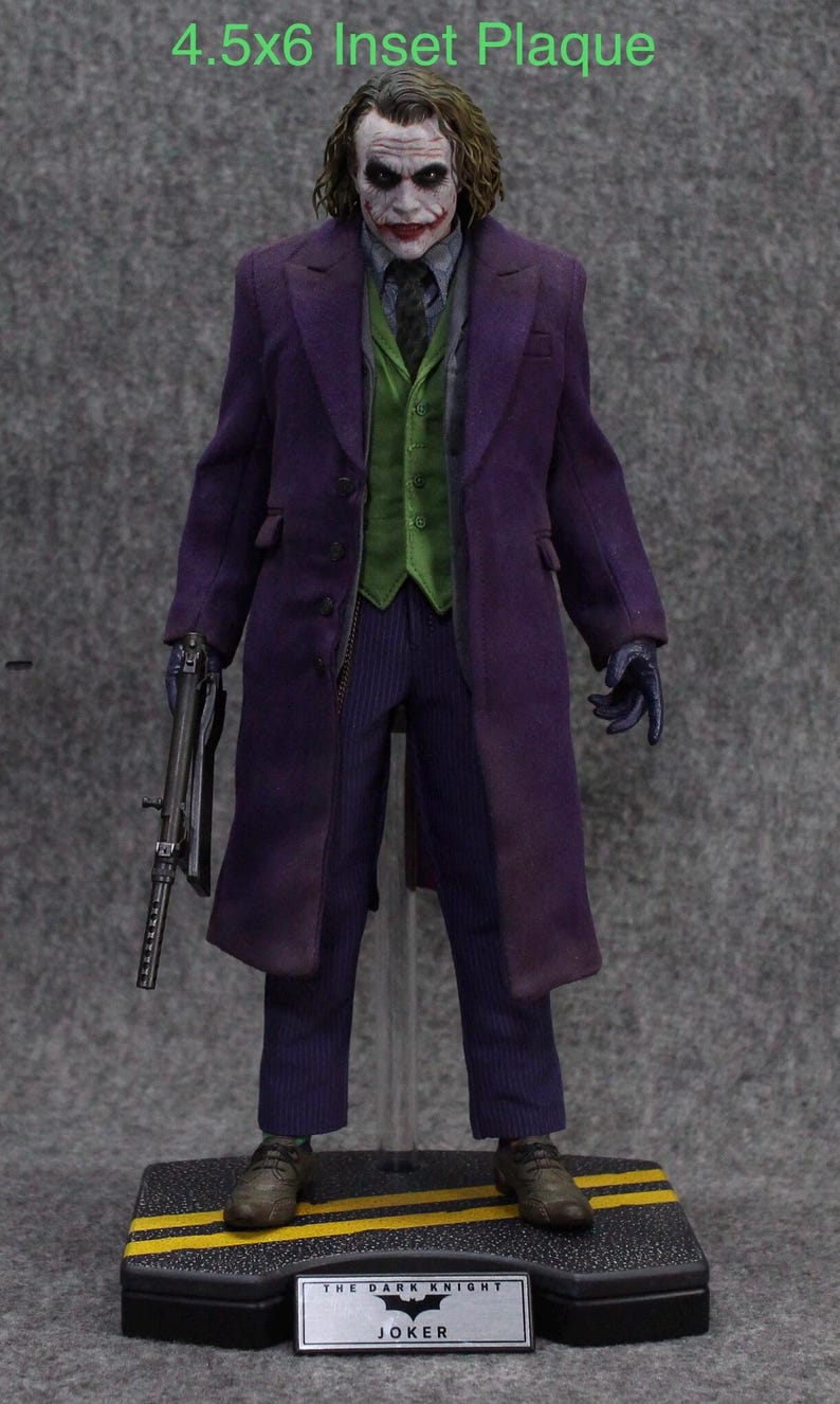 K&ouml;nnte beinhalten: Eine lila und gr&uuml;ne Joker-Actionfigur aus dem Film The Dark Knight. Die Figur h&auml;lt eine Waffe und steht auf einem schwarzen Sockel mit einem gelben Streifen. Der Sockel hat eine silberne Plakette mit der Aufschrift "The Dark Knight Joker".