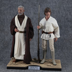 K&ouml;nnte beinhalten: Zwei Star Wars Actionfiguren, Obi-Wan Kenobi und Luke Skywalker, stehen auf einer W&uuml;stenbasis. Obi-Wan tr&auml;gt einen braunen Umhang &uuml;ber einer wei&szlig;en Tunika, w&auml;hrend Luke eine wei&szlig;e Tunika und Hose tr&auml;gt. Die Basis hat eine schwarze Plakette mit den Worten "MOVIE TITLE" und "CHARACTER NAME".