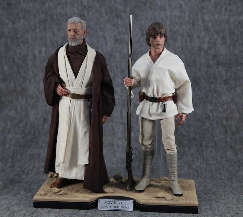 K&ouml;nnte beinhalten: Zwei Star Wars Actionfiguren, Obi-Wan Kenobi und Luke Skywalker, stehen auf einer W&uuml;stenbasis. Obi-Wan tr&auml;gt einen braunen Umhang &uuml;ber einer wei&szlig;en Tunika, w&auml;hrend Luke eine wei&szlig;e Tunika und Hose tr&auml;gt. Die Basis hat eine schwarze Plakette mit den Worten "MOVIE TITLE" und "CHARACTER NAME".