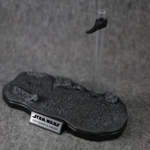 Puede incluir: Una base de exhibici&oacute;n gris para figuras de acci&oacute;n con un borde negro y un soporte transparente. La base tiene una superficie texturizada y una placa de identificaci&oacute;n met&aacute;lica que dice "STAR WARS DIN DJARIN & GROGU".