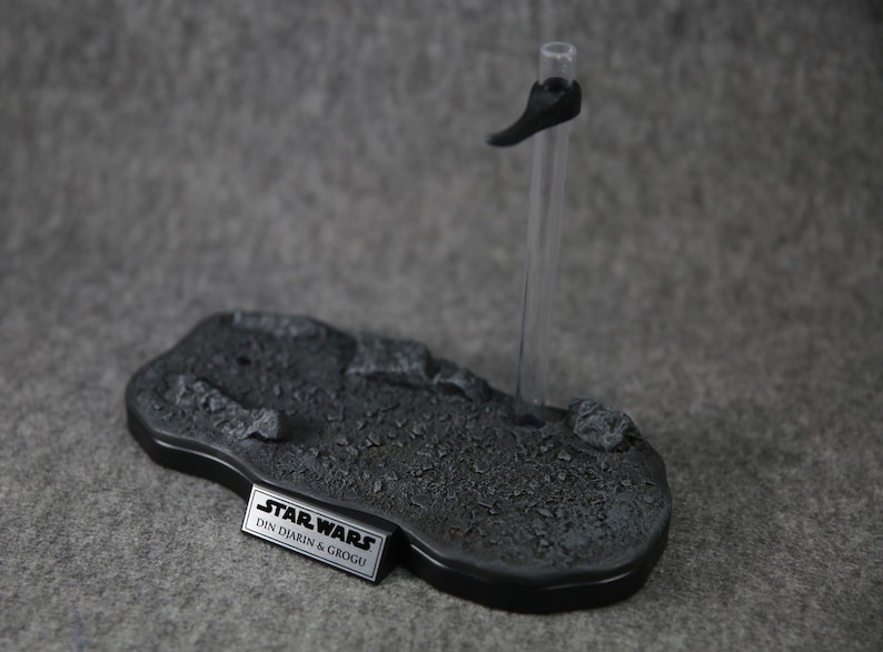 Puede incluir: Una base de exhibici&oacute;n gris para figuras de acci&oacute;n con un borde negro y un soporte transparente. La base tiene una superficie texturizada y una placa de identificaci&oacute;n met&aacute;lica que dice "STAR WARS DIN DJARIN & GROGU".