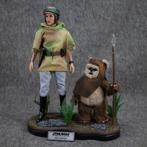 May include: A Star Wars action figure set featuring Princess Leia in a camouflage outfit and a Wicket action figure holding a spear. The figures are standing on a base with a green and brown background. The base has a label that reads "Star Wars Return of the Jedi Leia & Wicket".