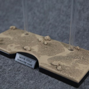 K&ouml;nnte beinhalten: Ein beigefarbener Displayst&auml;nder mit einer W&uuml;stentextur und zwei klaren Acrylst&auml;ben. Der St&auml;nder hat eine silberne Plakette mit dem Text "MOVIE TITLE CHARACTER NAME".