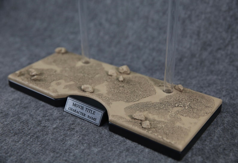 K&ouml;nnte beinhalten: Ein beigefarbener Displayst&auml;nder mit einer W&uuml;stentextur und zwei klaren Acrylst&auml;ben. Der St&auml;nder hat eine silberne Plakette mit dem Text "MOVIE TITLE CHARACTER NAME".