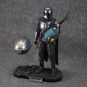 Puede incluir: Figura de acci&oacute;n detallada de The Mandalorian, con armadura plateada, sosteniendo a Grogu envuelto en una manta azul. Un casco desmontado descansa cerca. La base presenta el logo de Star Wars y el texto "Din Djarin & Grogu."