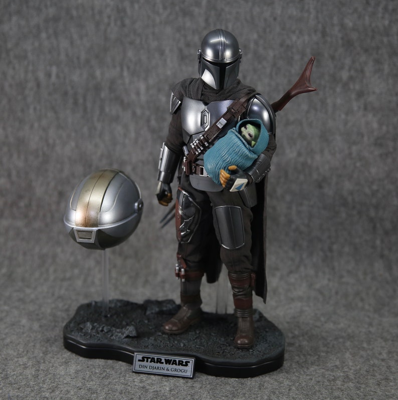 Puede incluir: Figura de acci&oacute;n detallada de The Mandalorian, con armadura plateada, sosteniendo a Grogu envuelto en una manta azul. Un casco desmontado descansa cerca. La base presenta el logo de Star Wars y el texto "Din Djarin & Grogu."