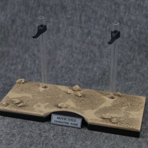 K&ouml;nnte beinhalten: Ein brauner und schwarzer Pr&auml;sentationsst&auml;nder mit zwei klaren Acryl-Stangen und schwarzen Kunststoffhaltern. Der St&auml;nder hat eine strukturierte Oberfl&auml;che, die an Sand erinnert, und ein Etikett mit der Aufschrift "MOVIE TITLE CHARACTER NAME".