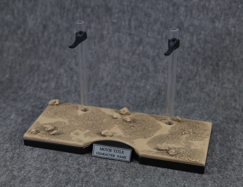 K&ouml;nnte beinhalten: Ein brauner und schwarzer Pr&auml;sentationsst&auml;nder mit zwei klaren Acryl-Stangen und schwarzen Kunststoffhaltern. Der St&auml;nder hat eine strukturierte Oberfl&auml;che, die an Sand erinnert, und ein Etikett mit der Aufschrift "MOVIE TITLE CHARACTER NAME".