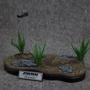 May include: A Star Wars diorama featuring Leia and Wicket from Return of the Jedi. The diorama is made of brown and green materials and features a small stand with a clear plastic rod. The diorama is set on a gray background.