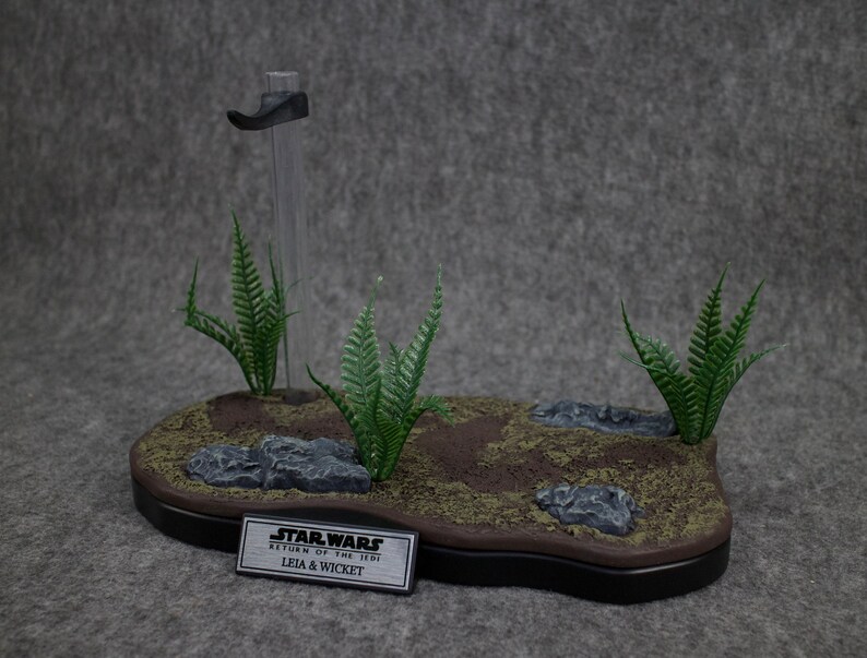 May include: A Star Wars diorama featuring Leia and Wicket from Return of the Jedi. The diorama is made of brown and green materials and features a small stand with a clear plastic rod. The diorama is set on a gray background.