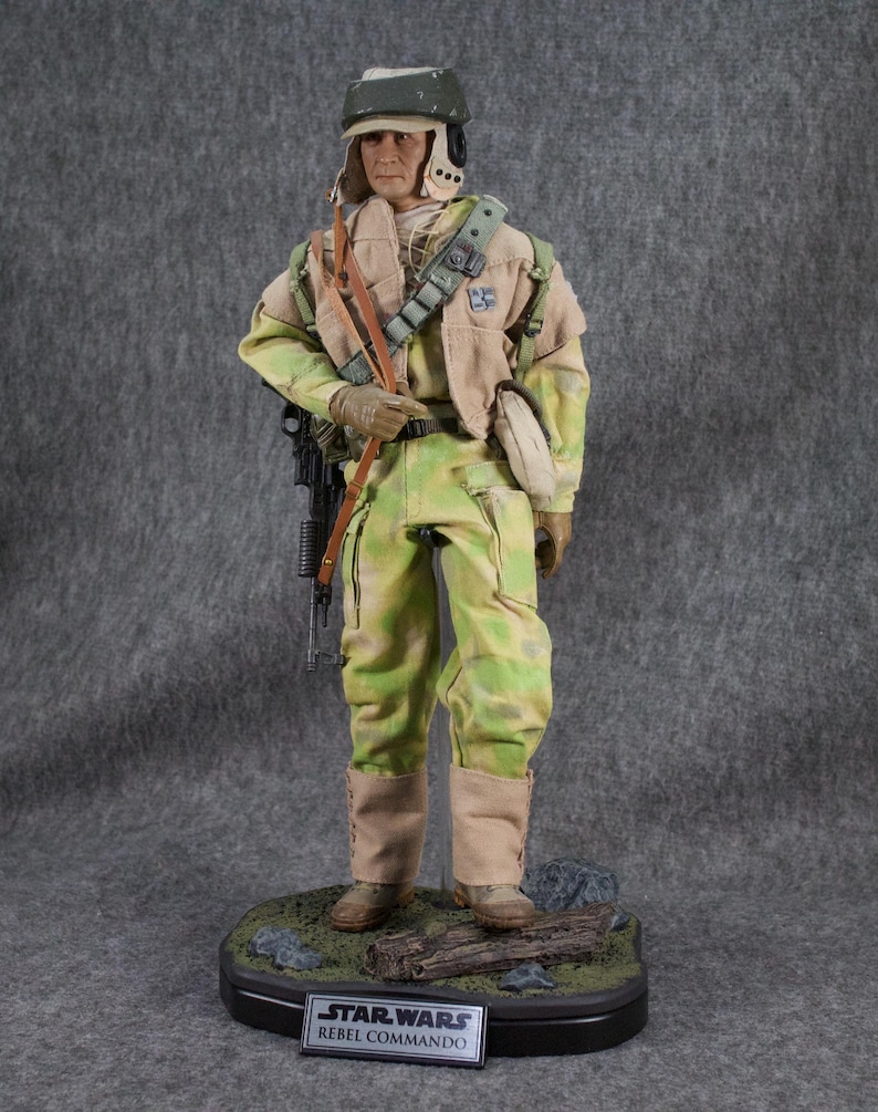 May include: A detailed action figure of a Rebel Commando from Star Wars. The figure is wearing a green and brown camouflage uniform, a helmet, and carrying a blaster rifle. The figure is standing on a base with a Star Wars logo and the words "Rebel Commando".