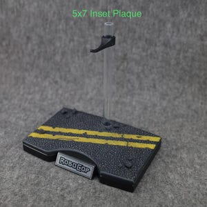 K&ouml;nnte beinhalten: Ein schwarzer Plastik-Actionfigurenst&auml;nder mit einer durchsichtigen Plastikstange und einem schwarzen Griff. Die Basis des St&auml;nders hat ein schwarzes und gelbes Stra&szlig;enmuster mit dem Schriftzug "RoboCop" auf einer silbernen Plakette.