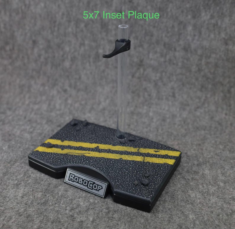 K&ouml;nnte beinhalten: Ein schwarzer Plastik-Actionfigurenst&auml;nder mit einer durchsichtigen Plastikstange und einem schwarzen Griff. Die Basis des St&auml;nders hat ein schwarzes und gelbes Stra&szlig;enmuster mit dem Schriftzug "RoboCop" auf einer silbernen Plakette.