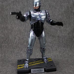 K&ouml;nnte beinhalten: Eine silberne RoboCop-Actionfigur mit einer schwarzen Pistole in der rechten Hand. Die Figur steht auf einem schwarzen Sockel mit gelben Linien und dem aufgedruckten Wort "RoboCop". Die Figur ist Teil einer 5x7 Zoll gro&szlig;en, eingelassenen Plakette.