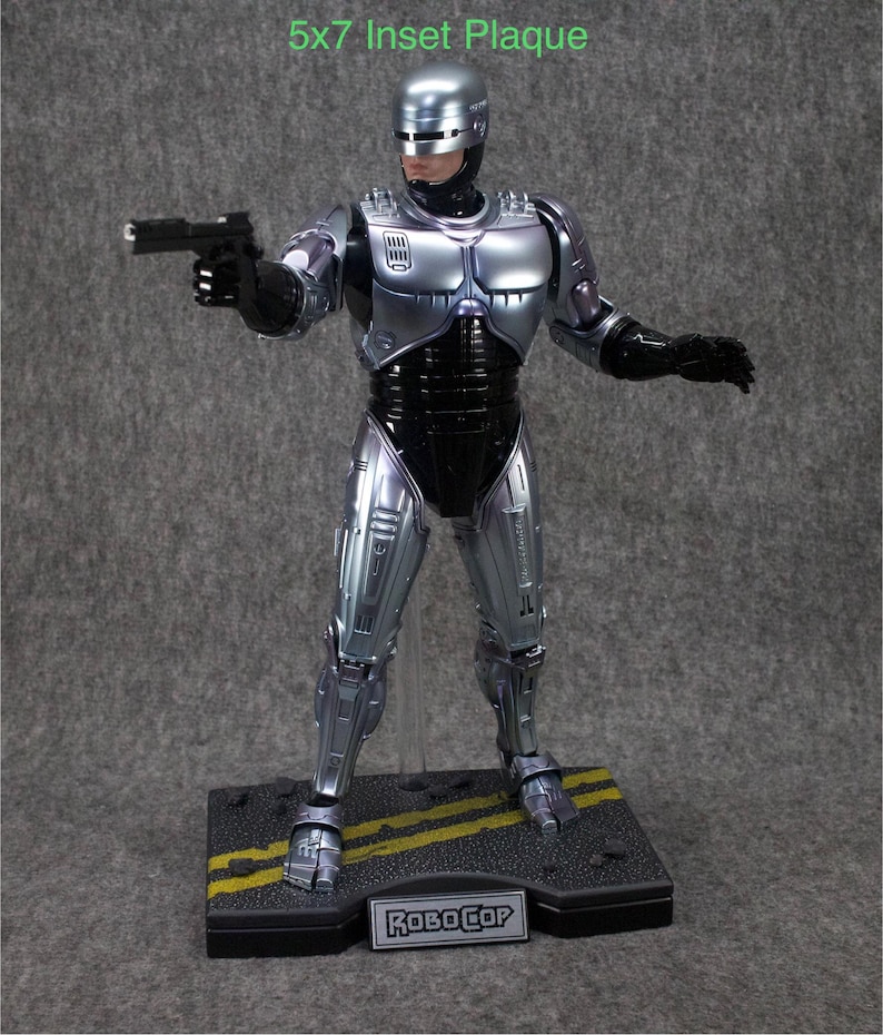 K&ouml;nnte beinhalten: Eine silberne RoboCop-Actionfigur mit einer schwarzen Pistole in der rechten Hand. Die Figur steht auf einem schwarzen Sockel mit gelben Linien und dem aufgedruckten Wort "RoboCop". Die Figur ist Teil einer 5x7 Zoll gro&szlig;en, eingelassenen Plakette.
