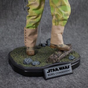 May include: A Star Wars Rebel Commando action figure standing on a base with a log and rocks. The figure is wearing a green and brown camouflage uniform with tan boots. The base has a black plaque with the words "Star Wars Rebel Commando".