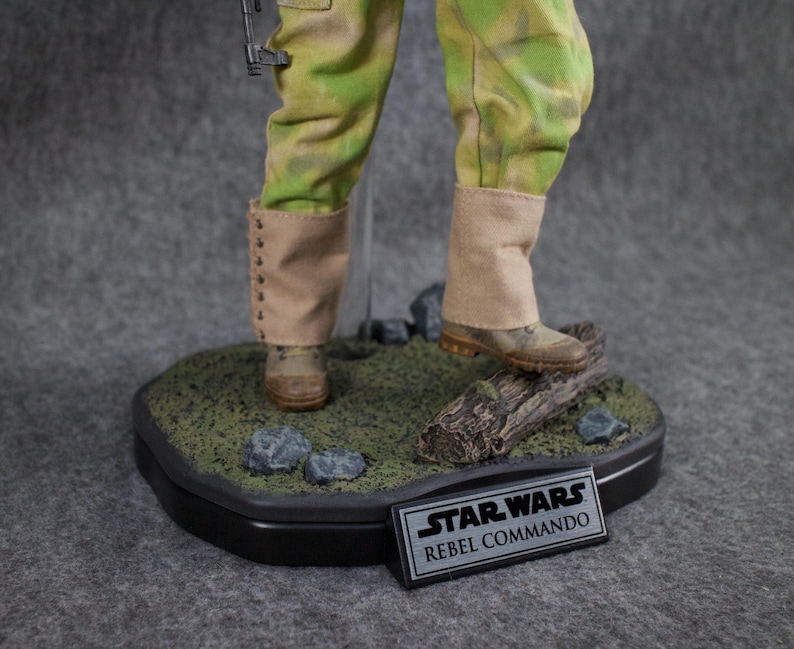 May include: A Star Wars Rebel Commando action figure standing on a base with a log and rocks. The figure is wearing a green and brown camouflage uniform with tan boots. The base has a black plaque with the words "Star Wars Rebel Commando".
