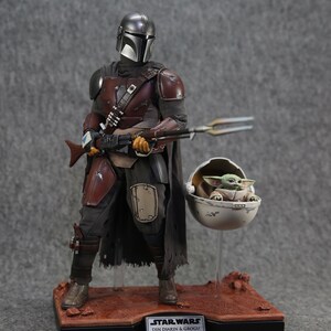 May include: A Star Wars action figure set featuring Din Djarin, also known as The Mandalorian, and Grogu, also known as Baby Yoda. The Mandalorian is wearing his signature armor and holding a blaster rifle. Grogu is sitting in a floating pod. The figures are displayed on a brown base with the text "STAR WARS DIN DJARIN & GROGU".