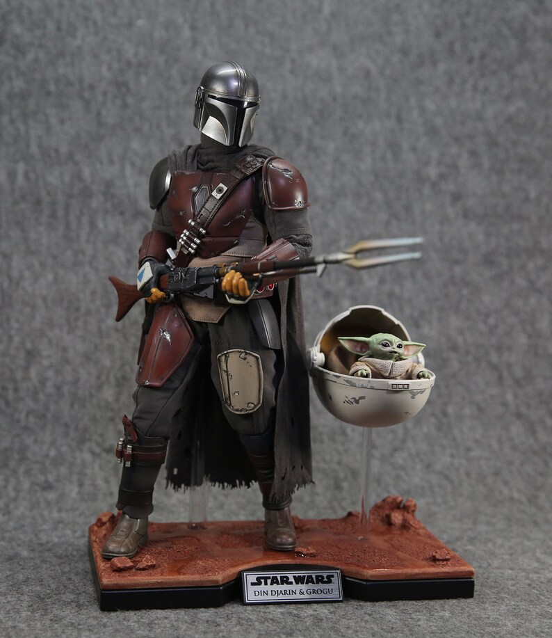 May include: A Star Wars action figure set featuring Din Djarin, also known as The Mandalorian, and Grogu, also known as Baby Yoda. The Mandalorian is wearing his signature armor and holding a blaster rifle. Grogu is sitting in a floating pod. The figures are displayed on a brown base with the text "STAR WARS DIN DJARIN & GROGU".