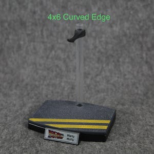 K&ouml;nnte beinhalten: Ein schwarzer Plastik-Actionfigurenst&auml;nder mit einem durchsichtigen Plastikstab und einer Basis mit abgerundeter Kante. Die Basis hat zwei gelbe Streifen und ein Etikett mit der Aufschrift "Zur&uuml;ck in die Zukunft" und "Marty McFly".