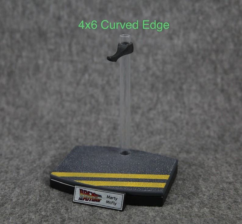 K&ouml;nnte beinhalten: Ein schwarzer Plastik-Actionfigurenst&auml;nder mit einem durchsichtigen Plastikstab und einer Basis mit abgerundeter Kante. Die Basis hat zwei gelbe Streifen und ein Etikett mit der Aufschrift "Zur&uuml;ck in die Zukunft" und "Marty McFly".