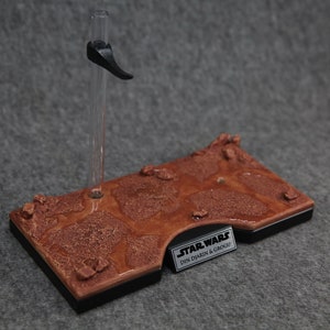 May include: A brown and black display stand with a textured surface that resembles a desert landscape. The stand has a clear plastic rod with a black clip at the top. The stand has a black label with the text "STAR WARS DIN DJARIN & GROGU".