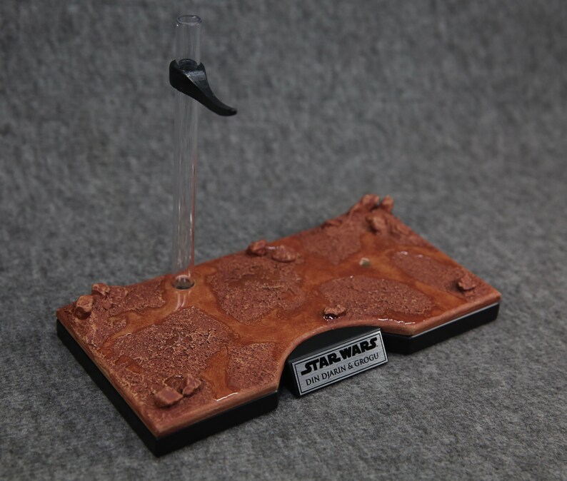 May include: A brown and black display stand with a textured surface that resembles a desert landscape. The stand has a clear plastic rod with a black clip at the top. The stand has a black label with the text "STAR WARS DIN DJARIN & GROGU".