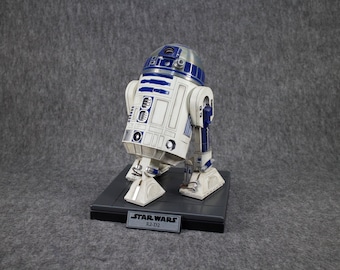 May include: A detailed R2-D2 figurine from Star Wars. The droid features a white body with blue accents and a silver head. It stands on a gray base with a Star Wars nameplate. The figurine is a collectible item.