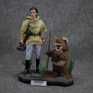May include: A Star Wars action figure set featuring Princess Leia and Wicket the Ewok. Leia is wearing a brown and green tunic with a belt and a blaster. Wicket is holding a spear and wearing a brown fur coat. The figures are standing on a black base with a green grass-like surface. The base has a silver plaque that reads "Star Wars Return of the Jedi Leia & Wicket".