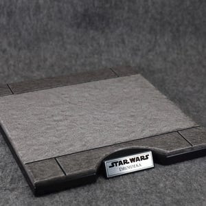 Puede incluir: Base de exhibici&oacute;n rectangular con una superficie gris texturizada y bordes grises oscuros. Una peque&ntilde;a placa plateada en la base dice "STAR WARS DROIDEKA". La base est&aacute; dise&ntilde;ada para exhibir objetos de colecci&oacute;n.