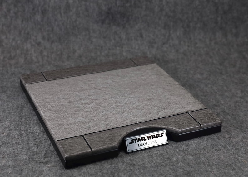 Puede incluir: Base de exhibici&oacute;n rectangular con una superficie gris texturizada y bordes grises oscuros. Una peque&ntilde;a placa plateada en la base dice "STAR WARS DROIDEKA". La base est&aacute; dise&ntilde;ada para exhibir objetos de colecci&oacute;n.
