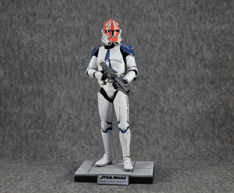 May include: A Star Wars 332nd Clone Trooper action figure. The figure is predominantly white with blue and orange accents. It is holding a black blaster rifle and stands on a gray base with the Star Wars logo and text.